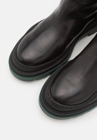 Bottes cheville en cuir noir à bouts arrondis, avec une semelle épaisse en caoutchouc vert et des coutures contrastantes le long des bords. Texture lisse dans l'ensemble.