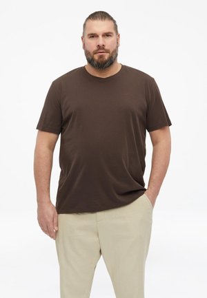 Mann mit Bart, der ein braunes kurzärmliges T-Shirt und beige Hose trägt, steht mit einer Hand in der Tasche vor einem einfarbigen weißen Hintergrund.