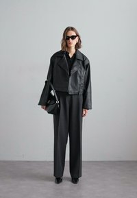 Giubbotto in pelle nera cropped sopra una camicia nera, abbinato a pantaloni larghi grigi. Gli accessori includono occhiali da sole neri e una borsa piccola.