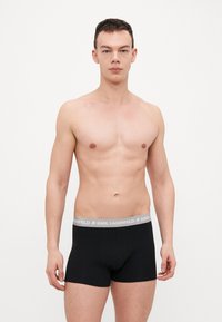 Melni boksera apakšveļas ar pelēku zīmola jostu. Materiāls izskatās mīksts un elastīgs. Modelis ir bez krekla, pozējot uz baltā fona.