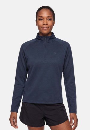 Femme portant un pull bleu marine à manches longues avec une fermeture éclair au quart et un short de sport noir, se tenant face à l'avant sur un fond uni.