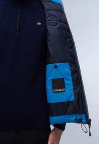 Marinblå stickad tröja under en blå och svart jacka. Jackan har quiltade detaljer, dragkedjestängning och en logotypmärke på den inre fickan.