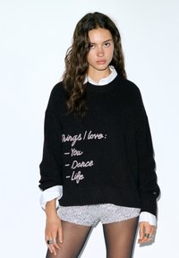 Pull noir en tricot avec une texture côtelée et une coupe surdimensionnée. Présente un texte brodé rose listant "You", "Dance" et "Life". Chemise blanche en dessous.