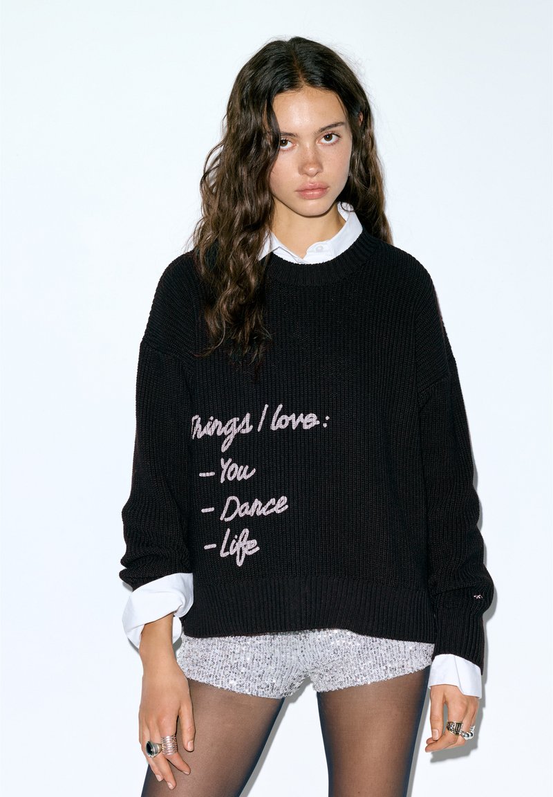 Czarny dziany sweter o prążkowanej fakturze i oversize'owym kroju. Na nim różowy haft z napisami "Ty", "Taniec" i "Życie". Pod spodem biała koszula.