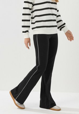 Person iført sorte bukser med hvide sidelister, beige sko og en hvid langærmet skjorte med sorte vandrette striber, stående på hvidt gulv.