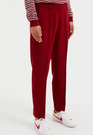 Person trägt rote maßgeschneiderte Hose, weiße Nike-Sneaker mit rotem Swoosh und ein rot-weiß gestreiftes Langarmshirt, steht auf einem weißen Boden.