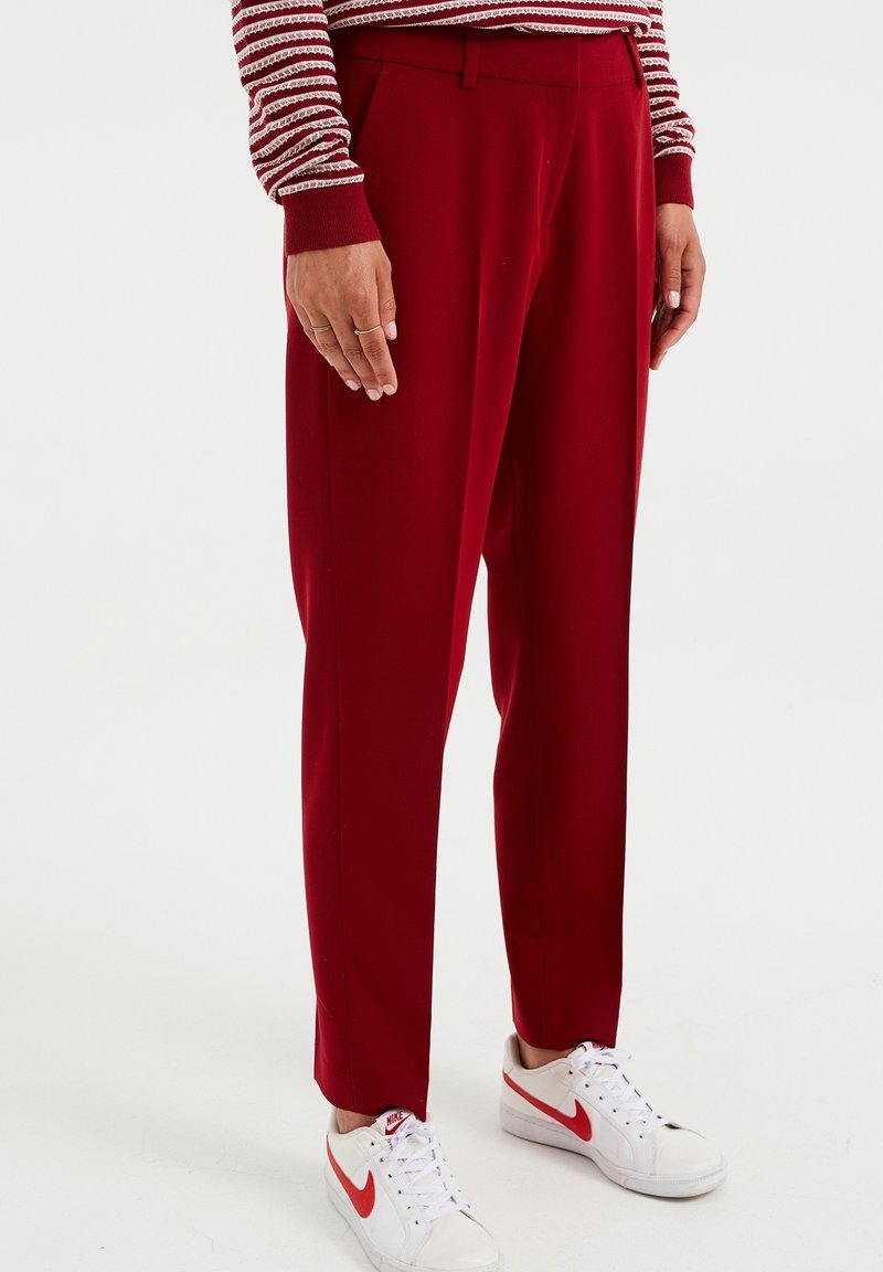 Personne portant un pantalon rouge sur mesure, des baskets blanches Nike avec le swoosh rouge, et un haut à manches longues rayé rouge et blanc, debout sur un sol blanc.