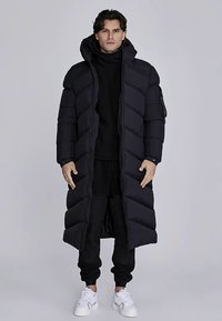 Lang sort dynejakke med quiltet mønster, udstyret med hætte og lynlåslukning foran, båret over en sort rullekrave og bukser, kombineret med hvide sneakers.