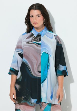 Kvinde med langt mørkt hår iført en løs, multifarvet skjorte med abstrakt mønster og matchende bukser, stående mod en hvid baggrund.