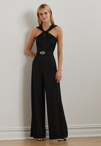 Svart jumpsuit med halterneck och snurrad design. Har en vida ben-silhuett, bältad midja med en dekorativ spänne, slät textur.