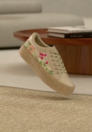 Zapatilla deportiva con plataforma beige, bordado floral y suela de yute trenzado, mostrada en el aire sobre una alfombra beige y una mesa de café de madera.