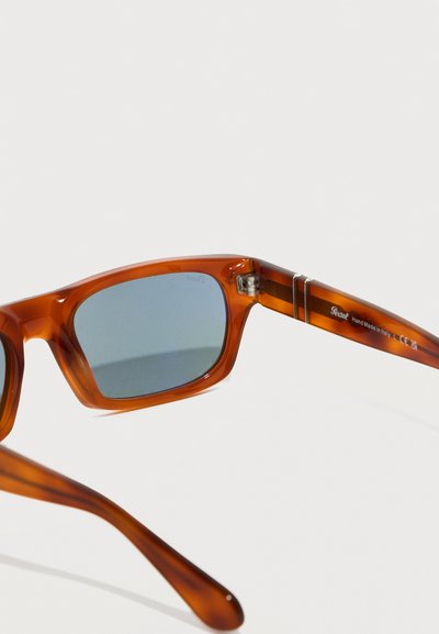 Καφέ ημιδιαφανή ορθογώνια γυαλιά ηλίου με μπλε φακούς, που παρουσιάζουν λεπτομέρεια άρθρωσης και το κείμενο "Κατασκευασμένα στο χέρι στην Ιταλία" στο μπράτσο.