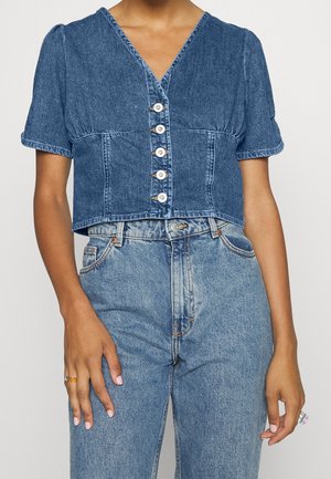 Kobieta ubrana w krótki, zapinany na guziki, niebieski denimowy top z krótkim rękawem, zestawiony z wysokim stanem niebieskich jeansów.