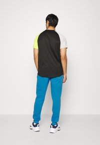 Muž s obličejem otočeným k odvrácené straně, oblečený v tričku s barevným blokem, které má černé, neonově žluté a bílé rukávy, modrých teplácích a bílých teniskách na jednoduchém pozadí.