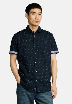 Man wearing a navy short-sleeve button-up shirt with small white dots and rolled cuffs, paired with blue jeans, standing against white background.