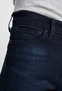 Tmavě modré džínové kalhoty s hladkou texturou, s klasickým pětikapsovým designem a středně vysokým páskem.