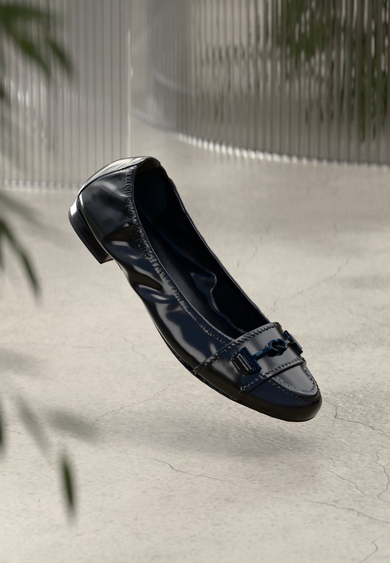 Mocassin plat en cuir noir brillant avec côtés élastiqués et embout métallique décoratif sur le dessus, présenté sur un fond neutre.