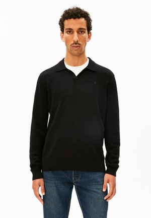 Mann mit kurzen lockigen Haaren und Schnurrbart, der ein schwarzes Langarm-Poloshirt über einem weißen Unterhemd und blauen Jeans trägt, steht vor weißem Hintergrund.