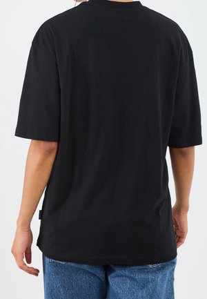Osoba ubrana w jednolity czarny t-shirt z krótkim rękawem i niebieskie jeansy, pokazana od tyłu na białym tle.