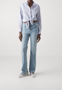 Gestreepte, lichtblauwe knoopshirt geknoopt in de taille; gecombineerd met lichtblauwe uitlopende jeans en grijze sneakers. Minimalistische esthetiek, casual pasvorm.