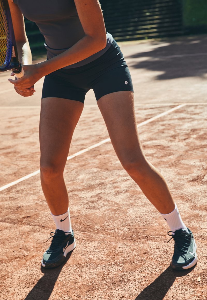 Juodi sportiniai šortai su mažu logotipu, dėvimi su pilku marškinėliu ir juodais Nike batais. Balti sportiniai kojinės ant molio teniso korto.
