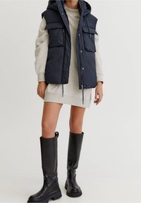 Femme portant une robe sweat-shirt grise, un gilet sans manches noir matelassé avec des poches et des bottes en cuir noires montant jusqu'aux genoux, debout sur un fond blanc.