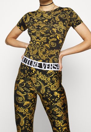 Žena oblečená v černém a zlatém topu na tělo s paisley vzorem a legínách s bílým pasem s nápisem "COUTURE VERSACE."