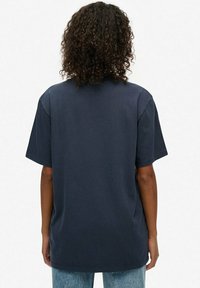 Osoba z kręconymi włosami, ubrana w prostą, ciemnoniebieską, oversize'ową koszulkę oraz jasnoniebieskie dżinsy, stojąca tyłem na białym tle.