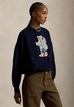 Unge med fletninger iført en navy sweatshirt med patchwork-teddybjørn design og brune bukser mod en ensfarvet baggrund.