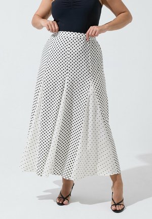 Femme portant une jupe midi blanche taille haute à pois noirs, associée à un haut noir sans manches et des sandales noires à talons.
