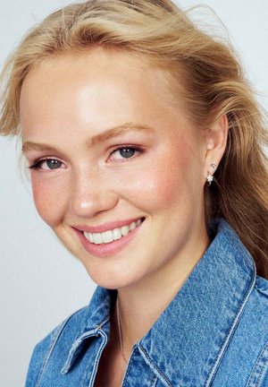Nærbillede af en smilende person med lyst blont hår iført en denimjakke, der viser fregnede kinder og små øreringe med blomstermotiv.