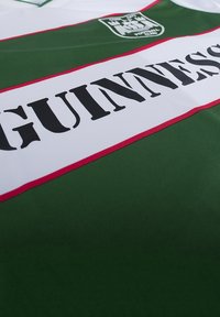 Groene en witte sporttrui met vetgedrukte zwarte "GUINNESS" tekst over de borst, verfraaid met rode strepen en een clubembleem op de schouder.