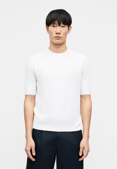 Pull en maille blanc à manches courtes avec une texture côtelée et un col rond, associé à un pantalon bleu marine foncé. Design simple et épuré.