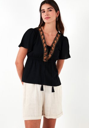 Zwarte blouse met korte pofmouwen, decoratieve oranje borduursels langs de halslijn en een verstelbare koord met kwastjes in de taille.