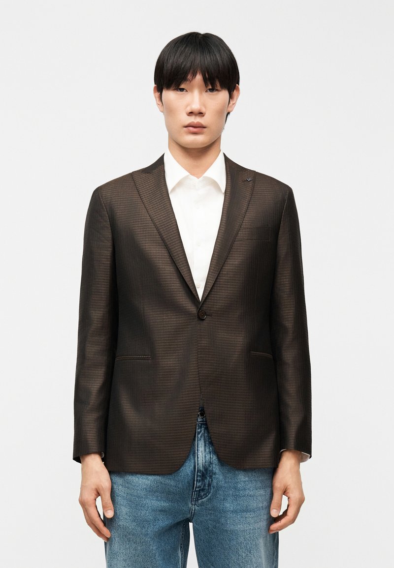 Homme portant un blazer texturé marron foncé sur une chemise blanche et un jean bleu, debout devant un fond clair uni.