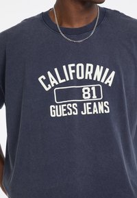 Granatowa bawełniana koszulka z białą grafiką, na której widnieje napis „CALIFORNIA 81 GUESS JEANS”. Klasyczny okrągły dekolt i luźny krój.