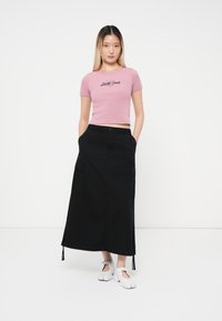 Ružičasti kratki top s kratkim rukavima i grafikom "Santa Cruz", uparen s crnom maksi suknjom koja ima džepove i vezice na rubovima. Bijele slip-on cipele.