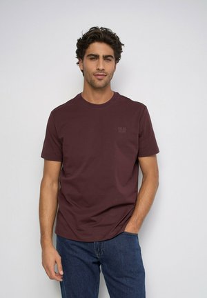 Hombre con cabello oscuro y ondulado, con una camiseta marrón lisa de manga corta y vaqueros azules, de pie con una mano en el bolsillo frente a una pared blanca.