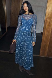 Mėlyna džinsinė maksi suknelė su ilgomis permatomomis rankovėmis, puošta raštuotu dizainu. Modelis laiko tamsiai rudą delninę ir dėvi tamsiai rudus batus.