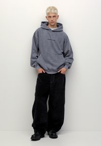 Hanorac supradimensionat gri cu buzunar frontal și text imprimat, purtat cu pantaloni largi din cord pentru bărbați negri și încălțăminte masivă neagră.