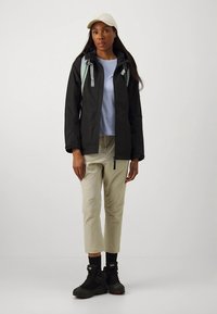 Čierna nepremokavá bunda, svetlomodrá košeľa, béžové krátke nohavice, čierne turistické topánky a béžová čiapka. Na bunde sú svetlomodré ramenné popruhy.