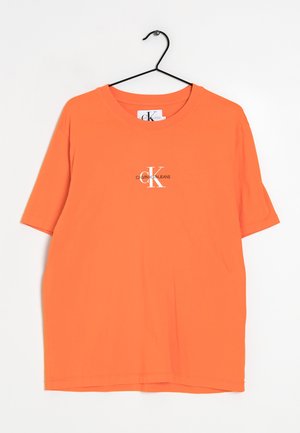 T-shirt en coton orange à manches courtes, avec un col rond et un logo imprimé en blanc sur la poitrine. Suspendu à un crochet noir.