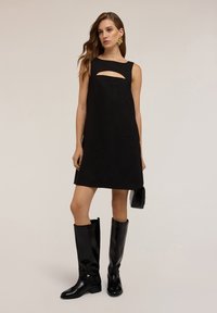 Vestido negro sin mangas con un detalle de corte, hecho de material similar al ante. Combinado con botas negras brillantes hasta la rodilla y un bolso pequeño.
