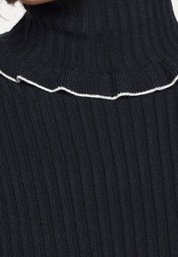 Svart ribbat turtleneck-tröja med en volangkrage i kontrasterande vit sömmar, som visar en närbild av halsområdet.