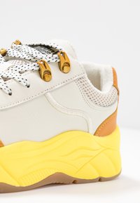 Sneaker featuring a white leather upper, mesh accents, yellow rubber sole, and gold eyelets for laces with textured details.