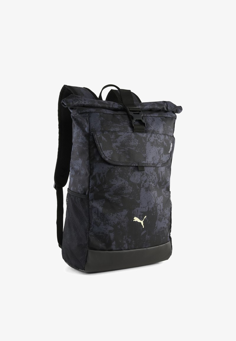 Zaino a motivi scuri con chiusura roll-top, spallacci neri, tasca laterale in rete e logo Puma dorato ben visibile sulla parte anteriore.