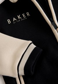Veste noire et beige avec poignets rayés, fermeture éclair et "BAKER by Ted Baker" imprimé à l'intérieur du col.