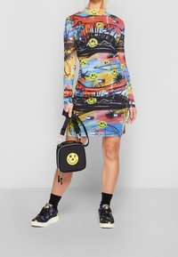 Mujer con un vestido colorido de cara sonriente y estampado abstracto, sosteniendo un bolso negro con una sonrisa amarilla y "NY", y usando zapatillas negras con sonrisa.