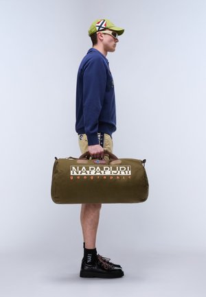 Mann mit grünem Cap, marineblauem Sweatshirt, beigen Shorts, schwarzen Schuhen, trägt eine olivgrüne Napapijri Reisetasche an den Griffen seitlich.