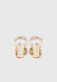 Gouden sandalen met hoge hakken, met een gesloten achterkant en enkelbandje. Beschikt over een gladde metalen afwerking en stevige blokhakken.
